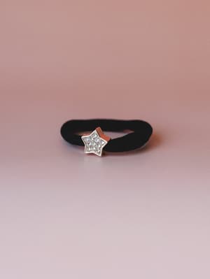 A black velvet hair tie with a pavé rhinestone star charm, shot solo against a soft dusty rose gradient — the kind of small thing that quietly makes a ponytail feel intentional.