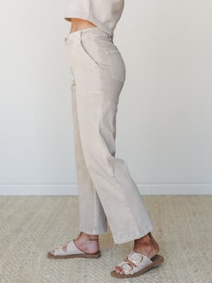 A side-profile shot showing wide-leg sand beige pants with patch pocket detailing, styled with a matching ribbed knit top and buckle slide sandals against a clean studio wall — effortless, tonal, and very much a 'Saturday errands feel good' outfit.