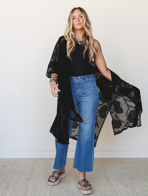 A relaxed full-body shot of a model layering a sheer black floral lace kimono over a black sleeveless top and cropped wide-leg jeans, styled with platform espadrille sandals and stacked jewelry — effortlessly boho with a cool-girl edge.