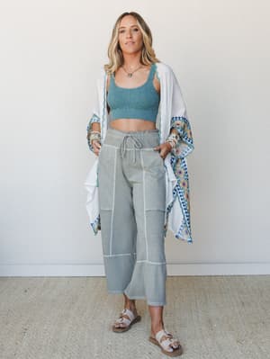 A breezy three-piece set styled on a model against a soft neutral wall — a crochet crop top in dusty teal pairs with relaxed wide-leg cropped pants in sage, layered under an open white kimono with colorful embroidered border trim.