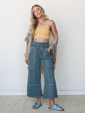 A breezy, sun-soaked layered look — wide-leg cropped pants in washed teal paired with a yellow crochet bralette top and a blush embroidered kimono draped loosely off the shoulders, finished with blue sandals and stacked bracelets.