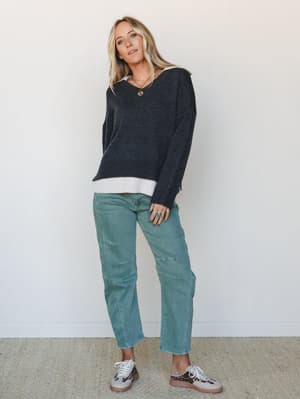 A relaxed, effortlessly layered look — a chunky charcoal v-neck sweater over a peeking white collared shirt, paired with sage green straight-leg jeans and leopard print sneakers, styled with gold and turquoise jewelry for that lived-in, personal-styl