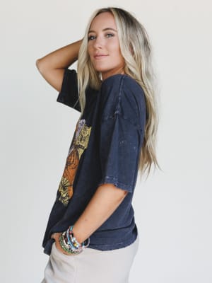 A woman with long blonde hair wears the On The Prowl Distressed Graphic Tee in charcoal and light pants. She stands sideways, looking at the camera with a slight smile, one arm bent to touch her head, colorful beaded bracelets on her wrist.