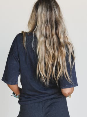 A person with long, wavy blonde hair is seen from behind, wearing the On The Prowl Distressed Graphic Tee in charcoal and dark pants. Bracelets adorn their wrist, and they stand against a plain, light background.