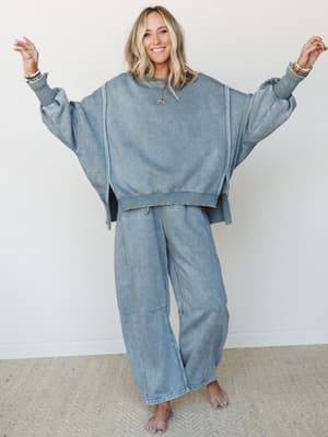 A smiling model wearing a matching acid-wash fleece set in a muted blue-grey — oversized cropped sweatshirt and wide-leg pants — arms spread wide like she's showing off the whole vibe, layered in beaded bracelets and a gold pendant necklace.