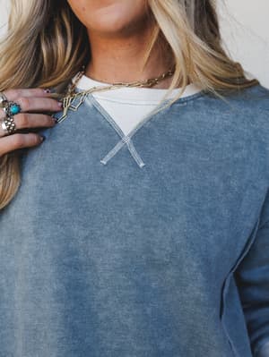 A close-up of a washed blue sweatshirt with a v-notch neckline detail and contrast cream layered collar, styled with a gold paperclip chain necklace and stacked boho rings — the kind of effortless, lived-in look that feels intentional without trying.