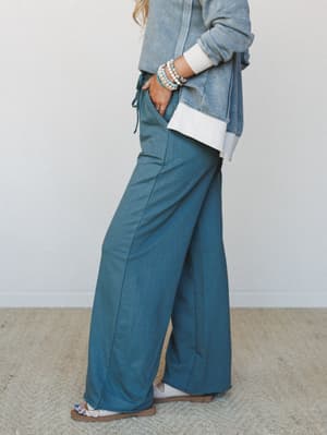 A side-profile shot focused on wide-leg pants in a muted teal-jade paired with a mineral-washed zip-up sweatshirt and layered white tee — the kind of effortless two-piece set that feels like a whole vibe without trying.