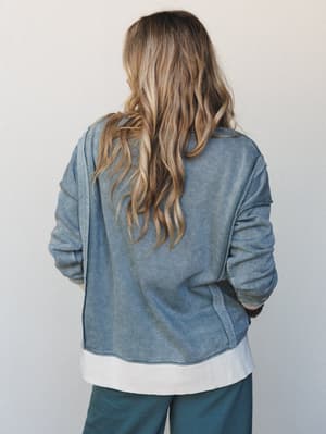 Shot from behind, a model in a washed indigo sweatshirt and teal wide-leg pants gives off that effortless 'just grabbed coffee and made it look intentional' energy — relaxed layering, soft tones, lived-in comfort.