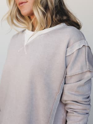 A close-up detail shot of a washed, lived-in sweatshirt in a muted ash-blue tone, layered over a white top — the tiered sleeve detail does the quiet, interesting work here.