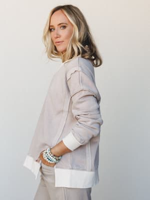 A model shot in three-quarter profile against a soft white wall, wearing a washed, oversized sweatshirt with exposed seam detailing and a white ribbed hem, layered over matching relaxed pants — the whole look reads effortlessly lived-in, finished wit