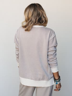 Shot from behind, a model wears a soft washed sweatshirt layered over a white long-sleeve tee, paired with relaxed wide-leg pants — the kind of effortless put-together that makes you want to stay in all day but also run errands.