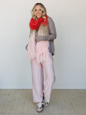A smiling blonde model wears a playful, mix-and-match three-piece set — a soft taupe long-sleeve top, pink-and-white striped wide-leg pants, and a color-blocked faux fur scarf in red, tan, and blush pink — paired with neutral sneakers for an effortle