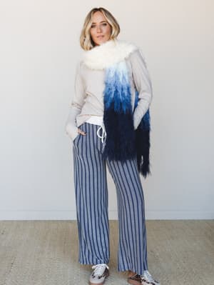 A relaxed full-body shot of a model styled in wide-leg striped pants, a soft long-sleeve top, and a statement ombre faux-fur scarf that goes from cream to deep navy — the kind of outfit that looks effortless but definitely took thought.