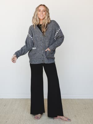 An oversized charcoal button-front cardigan with white whipstitch sleeve detail is layered over a black top and wide-leg black pants, finished with nude sandals — the kind of put-together-but-relaxed outfit that works just as hard at school pickup as