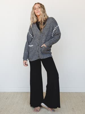 An oversized charcoal knit cardigan with white whipstitch detailing gets layered over a relaxed all-black set — wide-leg pants and a fitted tee — finished with nude strappy sandals for an effortlessly put-together everyday look.