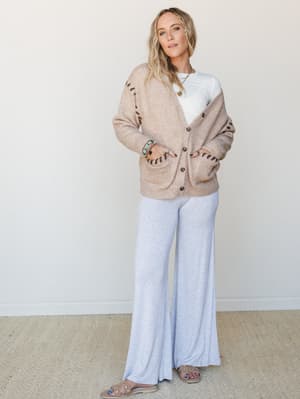 A relaxed, put-together set in warm neutrals — an oatmeal button-front cardigan with whipstitch detail layered over a white tee and flowy wide-leg pants in heather grey, styled with stacked turquoise bracelets and slide sandals for that effortlessly