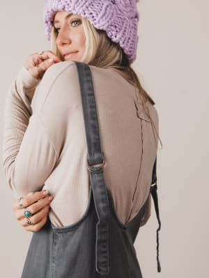 Person wearing a beige sweater and denim overalls with a purple knitted hat on a plain background