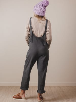 Person wearing a beige shirt, dark overalls, and a purple knitted hat against a plain wall.