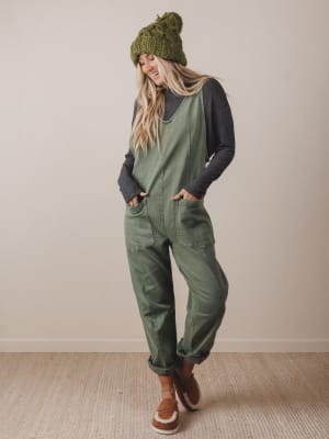 Person wearing a green knit beanie, gray long-sleeve shirt, and green jumpsuit with brown boots on a plain background.