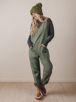 Person wearing a green knit beanie, gray long-sleeve shirt, and green jumpsuit with brown boots on a plain background.