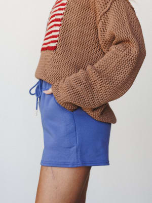A cropped side-angle shot showing a cobalt blue mini short with a drawstring waist, styled under a chunky open-knit camel sweater with a red and cream stripe detail peeking through — casual and a little playful, like she grabbed whatever felt good th