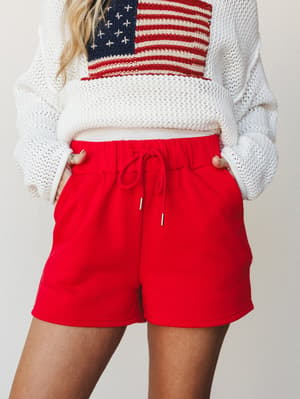 Red drawstring shorts paired with a cream knit sweater featuring an American flag graphic — a casual, patriotic combo that feels like a cookout you actually want to go to.