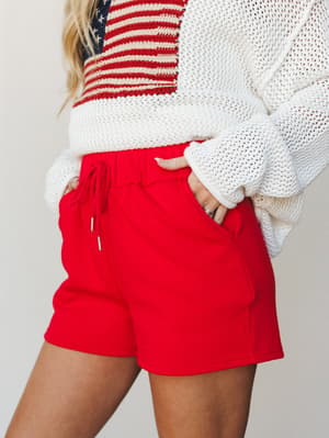 A close-up torso shot of red drawstring shorts paired with a cream open-knit sweater featuring an American flag graphic — casual, patriotic, and effortlessly summer-ready.