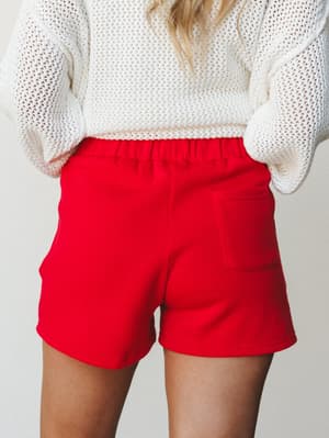 A back-view shot of bright ruby red pull-on shorts paired with a chunky cream open-knit sweater — casual, confident, and the kind of outfit that makes a Saturday feel intentional.
