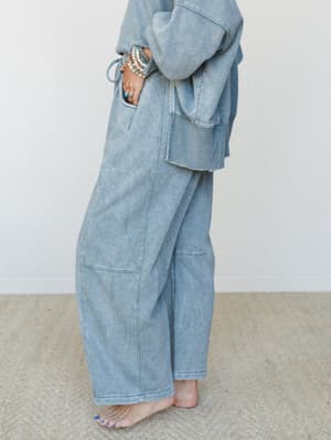 A side-angle shot of a matching acid-wash set in a soft ash blue-grey — wide-leg barrel sweatpants paired with an oversized cropped sweatshirt, styled barefoot with stacked turquoise and beaded bracelets for that effortless, lived-in feel.