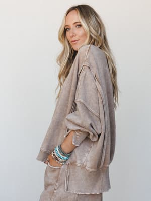 A relaxed, lived-in oversized sweatshirt in a washed camel tone shot in three-quarter profile — the kind of piece that looks equally at home on a coffee run or a cozy Sunday, styled with a stack of turquoise and beaded bracelets that do all the perso
