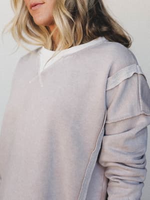 A close-up detail shot of a washed, lived-in sweatshirt in a soft muted lavender-grey, with layered ruffle sleeve detailing that gives it just enough personality to feel intentional — layered over a white tee and worn like a favorite.