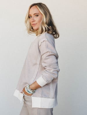 Shot in profile against a soft grey studio wall, she glances back over her shoulder in a relaxed, lived-in sweatshirt layered over a crisp white shirt, stacked bead bracelets adding just the right amount of personality.