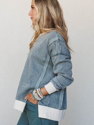 A relaxed side-profile shot of a model in a washed blue-grey sweatshirt with contrast cream cuffs and hem, styled with teal pants and stacked beaded bracelets — the kind of effortless layered look that feels equally at home on a coffee run or a slow