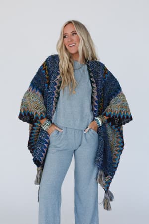A smiling woman with long blonde hair wears a light gray lounge set and the Desert Dreams Crochet Ruana - Cobalt, a colorful fringed knit shawl, adding an effortless boho touch. She stands with hands in her pockets against a plain background.