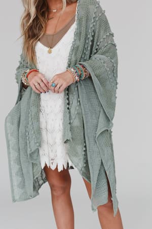 A woman wears a white lace dress paired with the Bali Embroidered Kimono in Sage, layered necklaces, and colorful beaded bracelets. Her wavy hair falls over her shoulder; only her torso and upper legs are visible.