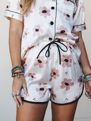 A close-cropped shot of a matching floral PJ short set in white with a retro daisy print — the black piping trim and ribbon drawstring give it that sweet-but-intentional finish, styled with a stack of beaded bracelets and silver rings that make it fe