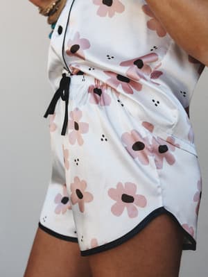 A close-up detail shot of satin floral pajama shorts in white with oversized retro daisy print in blush pink and black, styled with a matching top and tied black ribbon waistband — soft, playful, and made for slow mornings.
