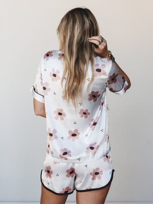 A back-facing shot of a satin floral pajama set — short-sleeve top and matching shorts in white with pink daisy print and black piping trim, styled with boho jewelry for a morning-after-but-make-it-cute feel.