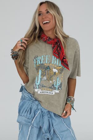 A smiling woman with long blonde hair wears a red bandana, boho-chic bracelets and rings, an oversized Fly Free Graphic Tee in Mocha, and a denim skirt, standing against a plain background.