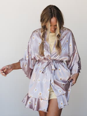 A satin daisy-print robe in soft lavender-pink worn over a yellow striped mini, belted at the waist with a self-tie — dreamy and a little retro, like getting ready is the whole event.