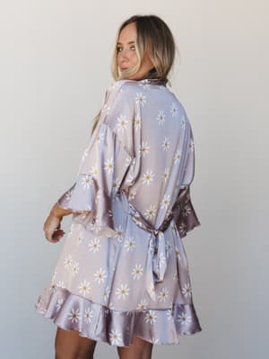A model glances back over her shoulder wearing a satin daisy-print robe with flutter sleeves and a tiered ruffle hem — the kind of piece that makes getting ready feel like the whole event.