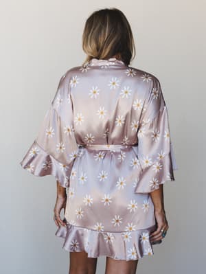 Shot from behind, a satin daisy-print mini robe in dusty mauve-pink with flutter sleeves and a ruffle hem — the kind of thing you'd wear to a bachelorette morning or just never want to take off.