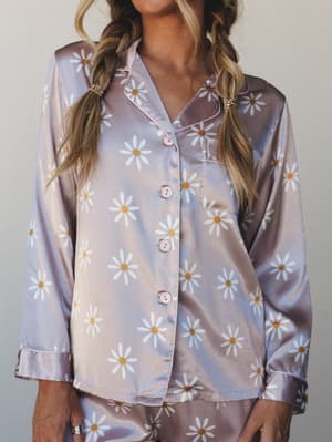 A satin pajama top in soft lavender-pink covered in cheerful white daisy prints — the kind of set that makes staying in feel like a whole personality.