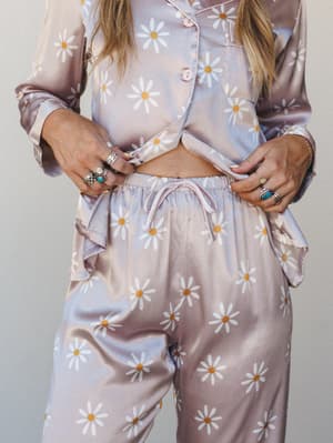 A daisy-print satin pajama set styled with stacked silver and turquoise rings — the kind of set that makes staying in feel like a whole personality.