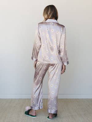 A model shot from behind showing a matching satin pajama set covered in an all-over daisy print — soft, dreamy, and the kind of thing you wear when staying in feels like an occasion.