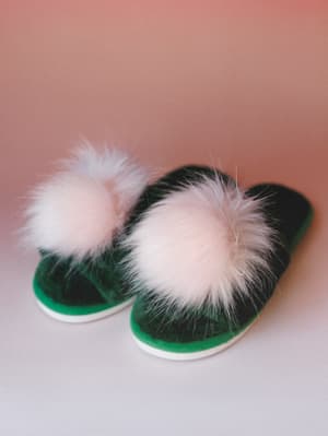 A pair of emerald green velvet slide slippers with oversized fluffy pom pom trim, shot against a soft dusty rose backdrop — the kind of cozy detail that makes staying in feel intentional.
