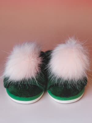 A pair of emerald green slippers with oversized blush pom-pom trim shot straight-on against a warm dusty rose backdrop — cozy and a little whimsical, like something you'd wear on a really good Saturday morning.