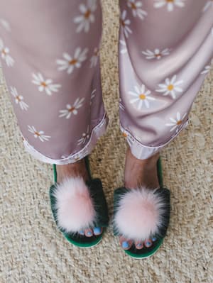 A top-down close-up of daisy-print satin wide-leg pants cuffed at the ankle, paired with green velvet slide slippers topped with oversized blush pom poms — standing on a woven jute rug with blue-painted toenails peeking through.