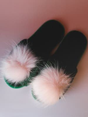 A pair of forest green open-toe slippers with oversized blush pink faux fur pom pom straps, shot from above on a soft pink-to-lavender gradient surface — cozy and a little whimsical.