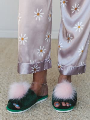 A close-up of satin daisy-print pajama pants with cuffed hems paired with forest green pom pom slippers — the kind of cozy, slightly absurd pairing that makes getting out of bed feel like a whole look.